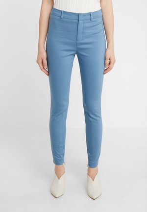 Femme portant un pantalon bleu clair coupe slim avec passants de ceinture, braguette zippée et fermetures éclair aux chevilles, associé à des chaussures pointues couleur crème.