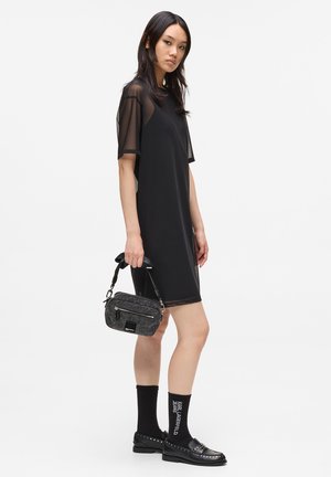 Giovane donna in un vestito nero corto con maniche trasparenti, tiene in mano una borsa grigia con texture, indossando calze nere e mocassini neri con borchie.