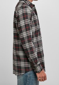 Camisa de franela a cuadros en blanco y negro con acentos rojos, de mangas largas, con cierre de botones en el frente y dobladillo redondeado, que presenta un cuello estándar.