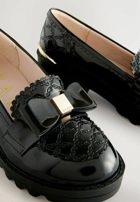 Černé lakované kožené loafers s texturovanou mašlí a zlatým čtvercovým detailem, a silnou, rýhovanou podrážkou.