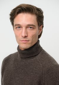 Brauner gerippter Rollkragenpullover aus strukturiertem Strickstoff mit hohem Kragen und taillierter Silhouette. Der Hintergrund ist neutral.