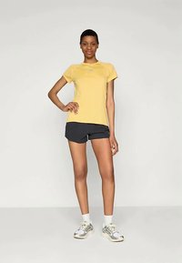 Keltainen urheilupaita lyhyillä hihoilla yhdistettynä tummiin shortseihin. Mallilla on myös päällään vaaleanharmaat lenkkarit ja valkoiset nilkkasukat.