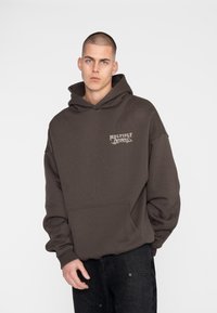 Felpa cappuccio marrone con tasca a marsupio e logo frontale. Il tessuto sembra morbido, con una vestibilità comoda e maniche lunghe. Indossata con jeans neri.