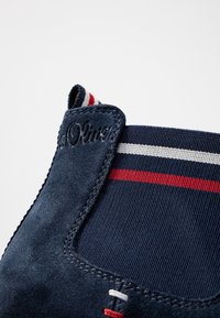 Botín de ante azul marino con paneles laterales elásticos con rayas rojas y blancas, costuras decorativas y logotipo grabado en la parte posterior.