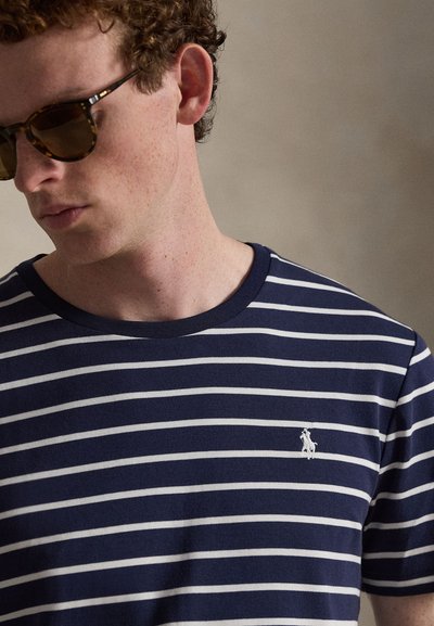 Νεαρός με σγουρά μαλλιά που φοράει γυαλιά ηλίου με σκελετό χελώνας και μπλε ναυτικό ριγέ μπλουζάκι με μικρό λευκό λογότυπο αλόγου.