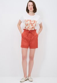 T-shirt bianco con stampa floreale rossa, abbinato a pantaloni corti color ruggine a vita alta con una cintura arricciata e cordoncino.