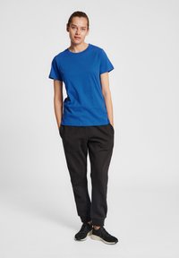 Blå bomulls t-shirt med korta ärmar, rund halsringning, kombinerad med svarta joggingbyxor och svarta sneakers, stående på en neutral bakgrund.