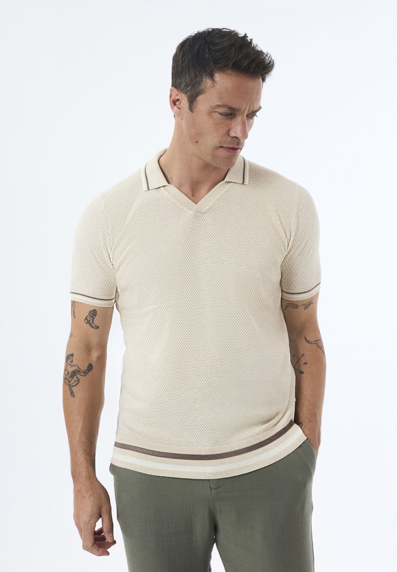 Camisa polo de manga corta beige con tejido texturizado, cuello en V y una franja marrón contrastante en el dobladillo. Se lleva con pantalones verdes.