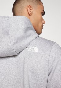 Sudadera gris de tela suave que presenta un logo blanco de "The North Face" en la parte posterior. El diseño incluye una capucha y un corte relajado.