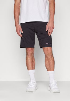 Pantalones cortos de algodón negros con un logo en la pierna izquierda, combinados con zapatillas y calcetines blancos. La camiseta es blanca y lisas.