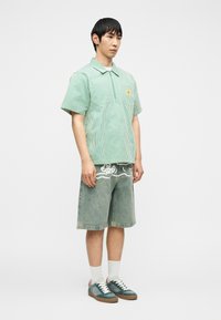 Νέος άνδρας με κοντομάνικο πουκάμισο με πράσινες ρίγες και φερμουάρ, τζιν σορτς με λευκή γραφική λεπτομέρεια, λευκές κάλτσες και πράσινα αθλητικά παπούτσια.