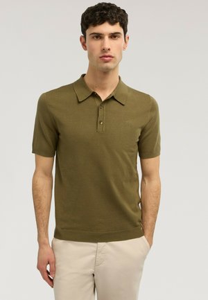 Mężczyzna w oliwkowej, krótkorękawowej koszulce polo i jasnobeżowych spodniach, stojący z jedną ręką w kieszeni na jednolitym tle.