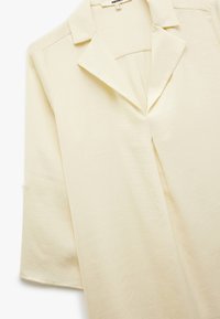 Cremefarbener Bluse mit einem eingekerbten Kragen, Dreiviertelärmeln und einem Schlitz vorne; glatte Textur und leichtes Material.