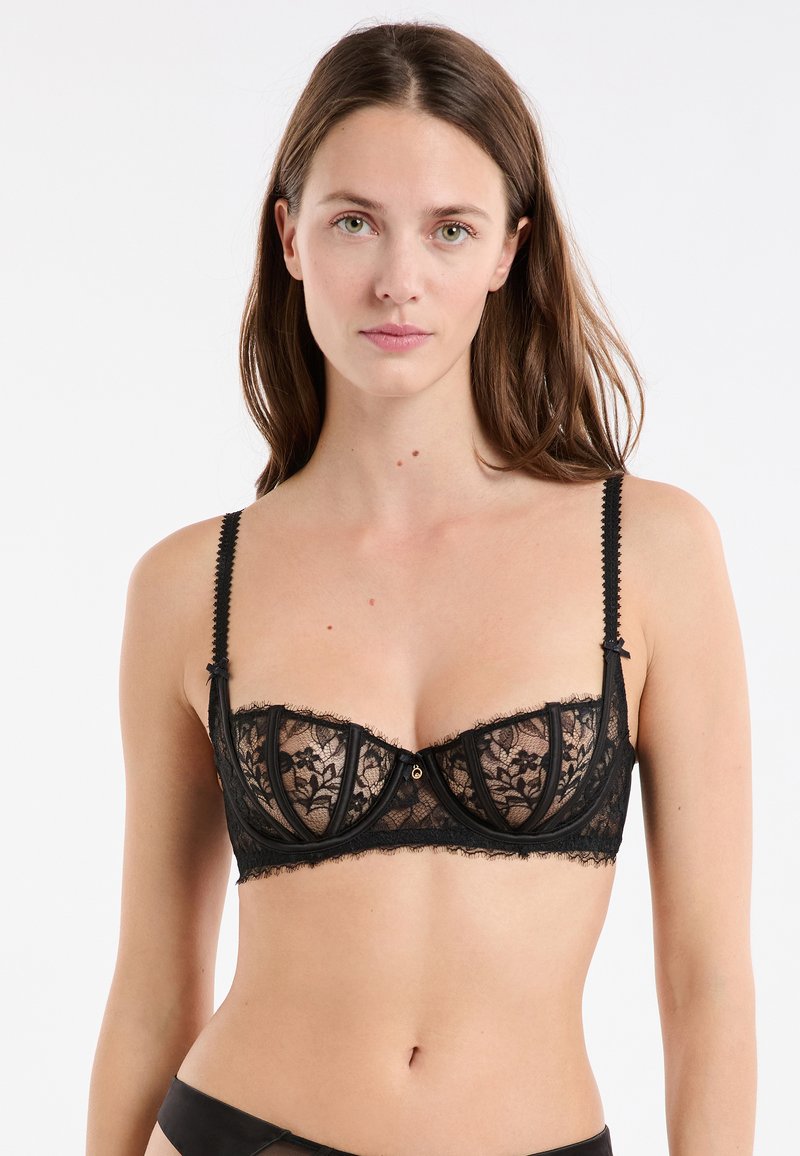 Joven mujer con cabello largo castaño, vistiendo un sujetador con aros de encaje negro, mirando hacia adelante contra un fondo blanco.