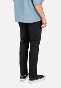 Hombre con pantalones negros, camisa azul y deportivas Converse azul marino, de pie con las manos relajadas a los lados sobre fondo blanco.