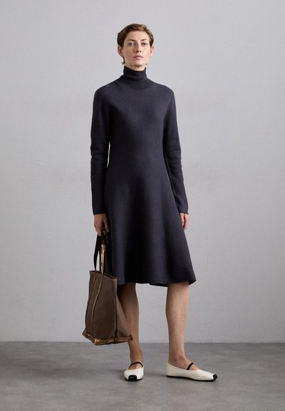 Graues Strickkleid mit hohem Kragen, langen Ärmeln und ausgestellt geschnittenem Saum. Dazu gehört eine braune Handtasche und weiße Ballerinas mit schwarzen Spitzen.