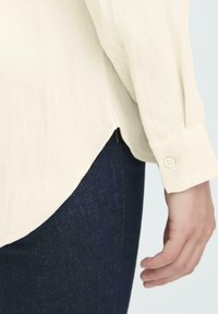 Light beige long-sleeve shirt cuff with button over dark blue denim jeans, showing side seam and a hand resting beside the hip.
