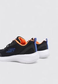 Schwarze Sportschuhe mit orangefarbener Innenausstattung, weißen Sohlen, blauem Logo und Fersenlaschen, nebeneinander auf einem einfarbigen Hintergrund platziert.