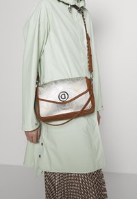 Metaliczna srebrna torebka na ramię z kwiatowym wzorem, brązowymi skórzanymi akcentami i okrągłym logo. Torebka ma regulowany pasek i zapięcie na klapkę.