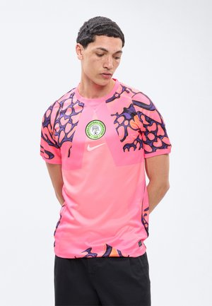 Νέος άνδρας κοιτάζει προς τα κάτω φορώντας μια ροζ φανέλα της Ομοσπονδίας Ποδοσφαίρου της Νιγηρίας με μαύρα και πορτοκαλί αφηρημένα μοτίβα στα μανίκια.