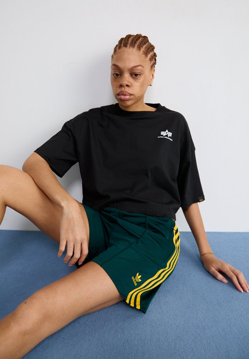 Svart bomullströja med rund hals och liten logga, tillsammans med gröna shorts med tre gula ränder och en adidas-emblem.