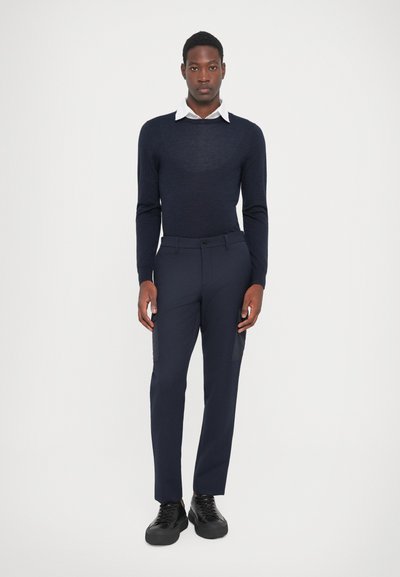 Pulover navy cu guler rotund, purtat peste o cămașă albă cu guler, asortat cu pantaloni navy și adidași negri. Textură netedă, design ajustat.
