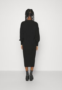 Robe noire en maille côtelée, coupe ample, manches longues, longueur midi, associée à des talons plateformes noirs épais et des chaussettes métalliques.