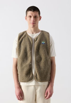 Jeune homme portant une veste sherpa beige sur un t-shirt et un pantalon crème, debout devant un fond clair uni.