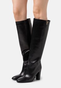 Bottes noires en cuir montantes avec un bout pointu, un talon carré et un accent doré en forme de "T" sur le côté extérieur, présentant une texture lisse et élégante.