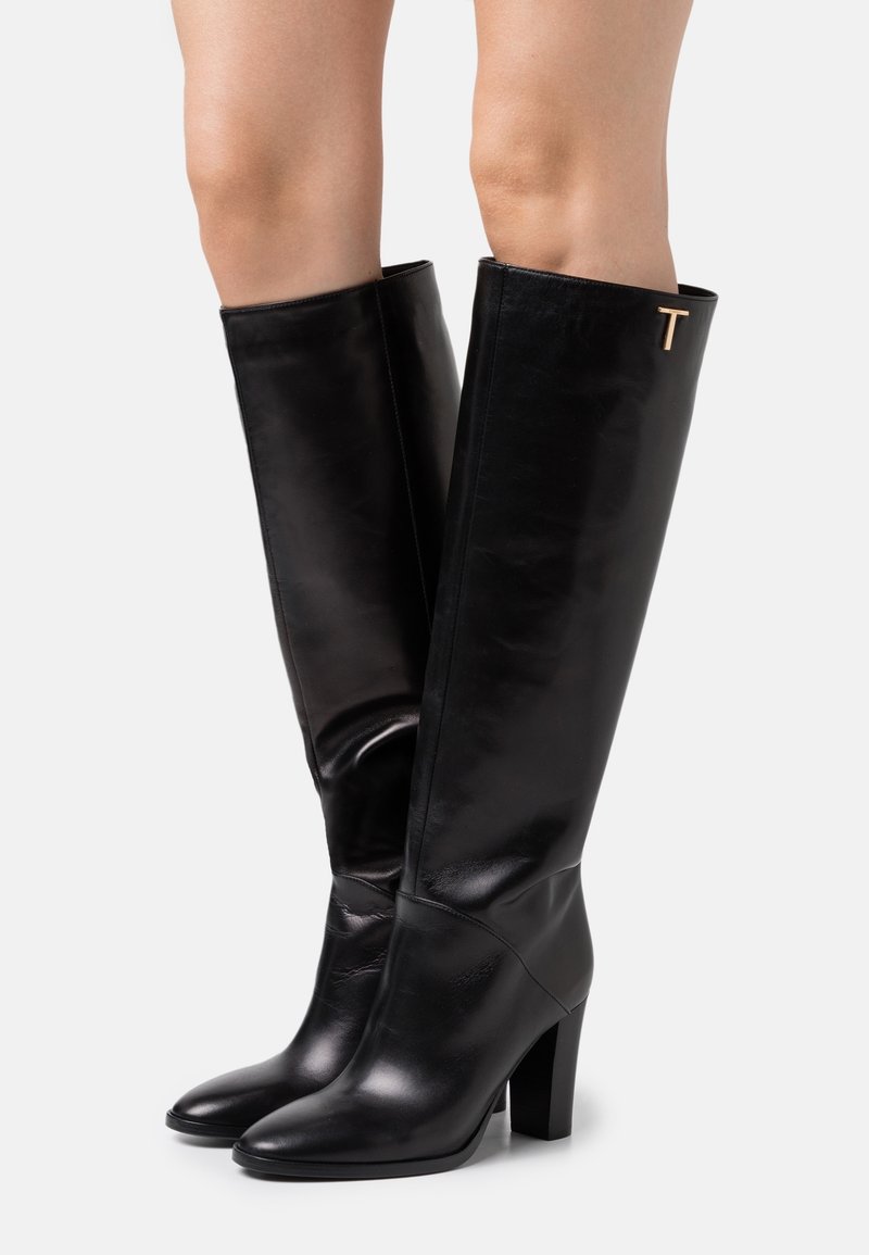 Bottes noires en cuir montantes avec un bout pointu, un talon carré et un accent doré en forme de "T" sur le côté extérieur, présentant une texture lisse et élégante.