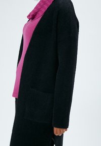 Cardigan nero peloso con tasca frontale, abbinato a un top rosa testurizzato con scollatura a cowl, visto di lato.