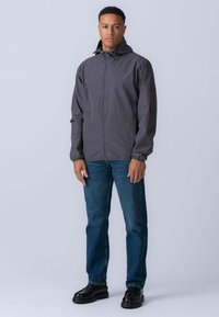 Grå, letvægtig jakke med hætte og frontlynlås. Båret over blå denimjeans og sorte sko, med et enkelt, funktionelt design.