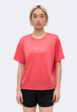 Femme aux cheveux blonds courts portant un t-shirt de sport corail à manches courtes et un short noir sur un fond gris uni.