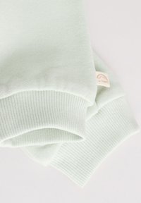 Poignets de sweat-shirt vert clair avec texture côtelée ; présente une petite étiquette crème avec un graphique arc-en-ciel indiquant "Je suis heureux."