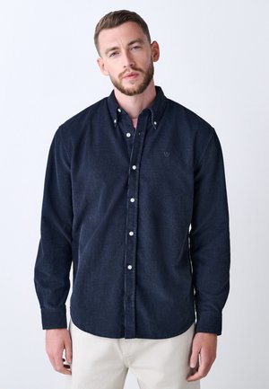 Mężczyzna o krótkich włosach i brodzie, ubraną w ciemnoniebieską koszulę zapinaną na guziki i jasne spodnie, stojący na białym tle.