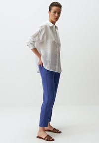 Witte linnen overhemd met lange mouwen, gecombineerd met blauwe op maat gemaakte broeken en bruine sandalen met bandjes. Eenvoudig ontwerp, casual uitstraling.