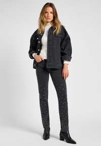 Jaqueta de ganga preta com picos decorativos nos ombros, combinada com jeans pretos com tachas dispersas. Modelo a usar botas de salto agudo.