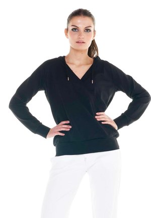 Une femme pose les mains sur les hanches, vêtue d’un sweat-shirt noir à capuche et d’un pantalon blanc, sur un fond blanc uni.