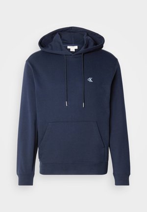 Tengerészkék kapucnis pulóver húzózsinórral, elülső kenguruzsebbel, bordázott mandzsettával és egy kis fehér logóval a mellkasán. Klasszikus dizájn.