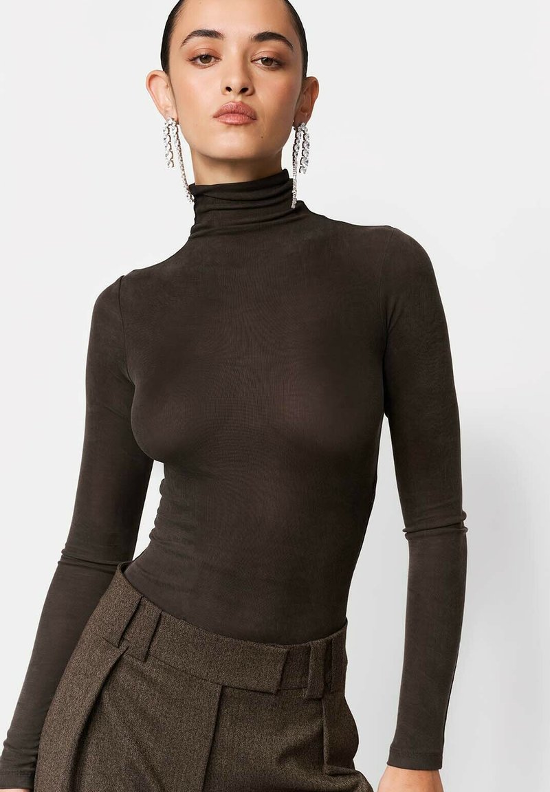 Brązowy dopasowany golf z długimi rękawami, wykonany z gładkiej tkaniny; zestawiony z wysokim stanem teksturowanych spodni i efektownymi kolczykami.