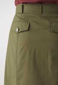 Oliwkowa spódnica o gładkiej teksturze, z dwoma bocznymi kieszeniami i detalem w postaci srebrnego zatrzasku. Dopasowany krój w talii.