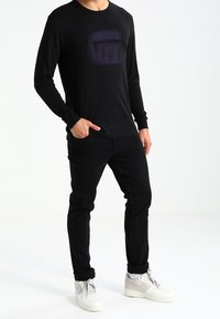 Musta pitkähihaista paitaa, jossa on violetti geometrinen logo rinnassa, yhdistetty mustiin housuihin ja valkoisiin lenkkikorkoihin. Silkkinen kankaan tekstuuri.