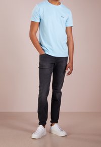 Muž v světle modrém tričku s krátkým rukávem, černých slim-fit džínách a bílých teniskách stojí s jednou rukou v kapse na jednobarevném pozadí.