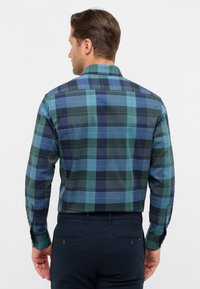 Uomo di spalle che indossa una camicia a righe blu e verde a maniche lunghe infilata in pantaloni blu navy, su uno sfondo semplice.
