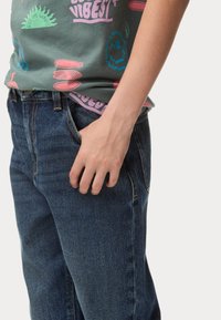 Jeans de denim azul con un ajuste clásico, que cuentan con bolsillos delanteros y una superficie sutilmente texturizada. Combinados con una camiseta gráfica.