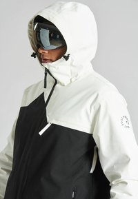 Chaqueta de esquí en blanco y negro con un bolsillo con cremallera, capucha ajustable y detalle del logo en la manga. Textura suave y diseño moderno.