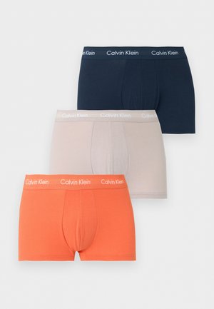 Atletický tmavší muž stojaci na boso v tmavomodrých Calvin Klein boxerkách na jednoduchom svetlom pozadí, jedna ruka na boku.