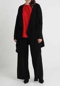 Cardigan nero aperto davanti su una maglietta rossa, abbinato a pantaloni neri a gamba larga. Tessuto morbido, vestibilità rilassata e design minimalista.