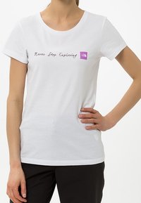 Bílý bavlněný tričko s kulatým výstřihem, krátkými rukávy a nápisem "Nikdy nepřestávejte prozkoumávat". Obsahuje fialovou nášivku s logem.