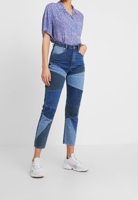 Žena v patchworkových modrých džínách, s fialovým vzorovaným tričkem s krátkým rukávem a bílými teniskami, stoj í na jednolitém bílém pozadí.